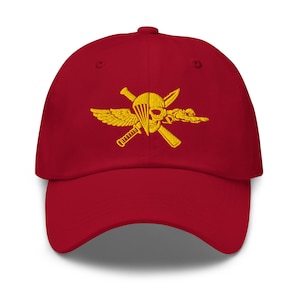 Può includere: Un cappellino da baseball rosso scuro con un emblema ricamato in giallo sul davanti. Il design mostra un teschio diviso verticalmente, combinato con un paracadute, un'ala singola e due pugnali incrociati, creando un grafico in stile militare.