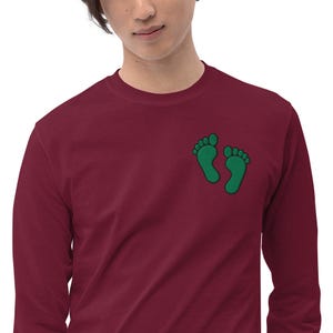 Può includere: Maglietta bordeaux a maniche lunghe con impronte di piedi verdi ricamate sul petto. La maglietta è a tinta unita e ha lo scollo tondo. Il design ricamato aggiunge un tocco giocoso al top casual.