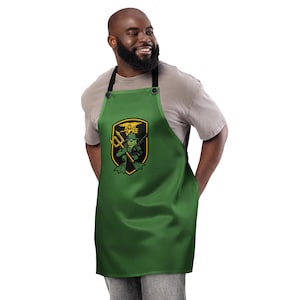 May include: Green apron with a black strap and a graphic of a frog with a trident and a rifle. The graphic is in a black and gold shield shape. The apron has two black buttons at the top.