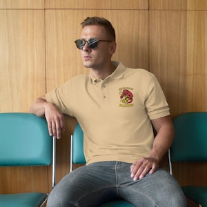 May include: A light tan polo shirt with a red devil graphic holding a soccer ball and the text "SUPER STOKED MARINES". The shirt has a classic collar and short sleeves. The person is wearing sunglasses and jeans.