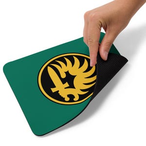 May include: A rectangular mouse pad with a dark green surface featuring a circular emblem in the center. The emblem displays a yellow sword and a stylized wing design against a black background. The underside of the mouse pad is black and textured.