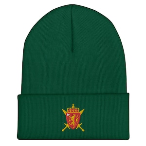 Könnte beinhalten: Eine grüne Strickmütze mit einem gestickten Wappen, das einen Löwen und Schwerter in Rot und Gold zeigt.