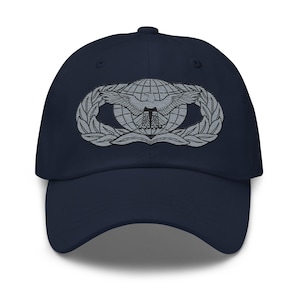Könnte beinhalten: Marineblaue Baseballkappe mit silbernem Emblem. Das Emblem zeigt einen Globus, einen Adler und einen Lorbeerkranz. Die Kappe hat einen gebogenen Schirm und einen Stoffriemenverschluss.