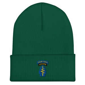 Puede incluir: Un gorro verde oscuro con un borde doblado. El gorro presenta un parche bordado con el texto "SPECIAL FORCES AIRBORNE" encima de un diseño de rayo. El gorro está hecho de un material de punto suave.