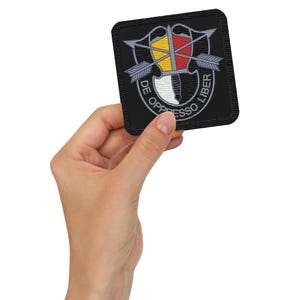 May include: A black embroidered patch with a silver shield, arrows, and a dagger. The text "DE OPPRESSO LIBER" is written around the shield.