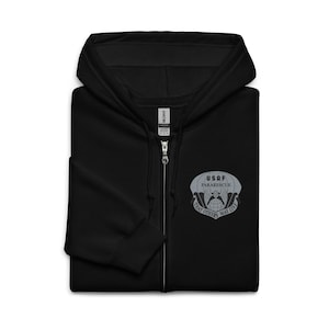 May include: Black zip-up hooded sweatshirt with a drawstring hood. The sweatshirt features a silver emblem on the left chest that reads "USAF Pararescue" with the text "That Others May Live" below. The zipper is silver.