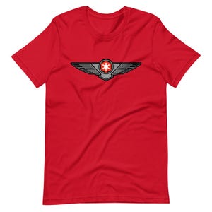 Puede incluir: Una camiseta roja con un gráfico de un emblema alado. El emblema tiene un diseño central de estrella, alas grises y negras y detalles en rojo. La camiseta está hecha de un material suave.