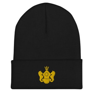 Puede incluir: Gorro de punto negro con un diseño bordado en oro de un casco de buceo flanqueado por dos caballitos de mar.