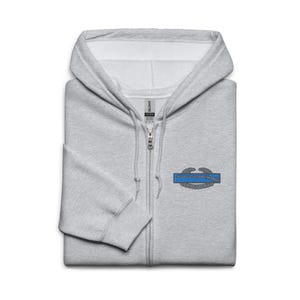 Può includere: Una felpa con cappuccio e zip grigio chiaro con una piccola toppa ricamata sul petto. La toppa presenta un rettangolo blu con testo e un disegno a corona argentata. La felpa ha una cerniera completa e un cappuccio.