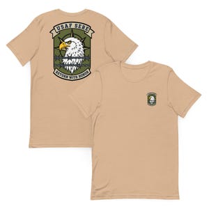 May include: Tan short-sleeve t-shirts. One shirt displays a large graphic on the back with a white eagle head, barbed wire, and text that reads "USAF SERE" and "RETURN WITH HONOR." The other shirt has a smaller version of the graphic on the front.