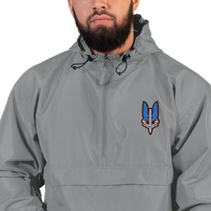 Puede incluir: Chaqueta cortavientos Champion gris con capucha y cierre de media cremallera. La chaqueta presenta un parche en el pecho con un emblema azul y rojo de alas, una espada y texto.