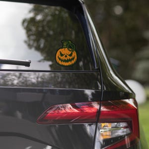May include: A Halloween sticker showing an orange jack-o'-lantern with a carved face, combined with a green grenade top. The sticker is attached to the rear window of a dark-coloured car, reflecting the surrounding environment.