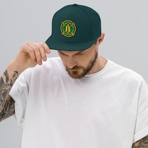 May include: A dark teal snapback cap featuring an embroidered circular patch on the front. The patch displays a yellow arrowhead pointing upward, centered over two crossed yellow arrows, all enclosed within a green circle with a yellow border.