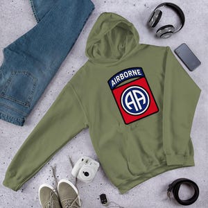 Könnte beinhalten: Ein olivgrüner Kapuzenpullover mit einem grafischen Aufnäher auf der Brust. Der Aufnäher zeigt den Text AIRBORNE in Weiß auf blauem Grund über einem roten Quadrat, das einen weißen Kreis mit den Buchstaben AA in Blau und Weiß enthält.