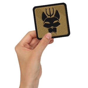 May include: Square embroidered patch with a black devil-like face design on a tan background, framed by a black border. The patch features a trident above the face and a jagged mouth. The patch is held in a hand.