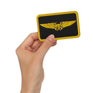 May include: A rectangular black patch with a gold embroidered emblem of wings and a shield. The patch has a gold border. The emblem is a symbol of aviation. The patch is held in a hand against a white background.