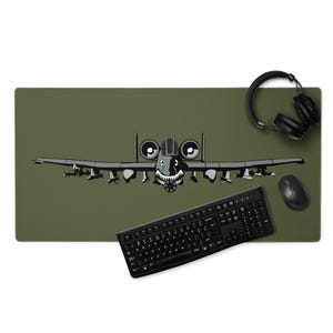 May include: An olive green desk mat featuring a cartoon illustration of a gray A-10 Thunderbolt II aircraft with a menacing grin. A black keyboard, mouse, and headphones are also on the mat.