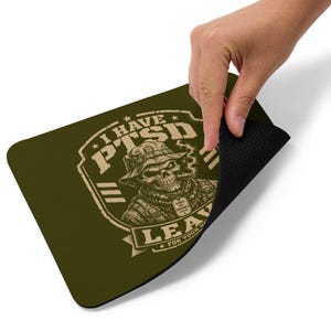 May include: An olive green rectangular mouse pad featuring a graphic of a skull wearing a military-style hat and dog tags. The text reads I HAVE PTSD LEAVE FOR YOUR SAFETY. The design is printed in a light tan color with a black rubberized underside.