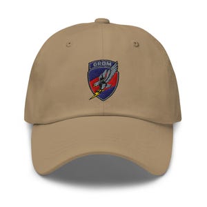 May include: A khaki baseball cap featuring an embroidered patch on the front. The patch is a shield shape with a blue and red background, displaying a grey eagle clutching a yellow lightning bolt, with the text GROM printed in white letters.
