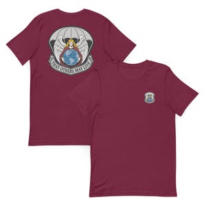 Può includere: T-shirt bordeaux con un disegno raffigurante un angelo che tiene in mano un globo sotto un paracadute, con la scritta "THAT OTHERS MAY LIVE". Una maglietta ha il disegno sul retro, l'altra sul davanti.