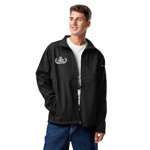 May include: A black softshell jacket featuring a white embroidered military-style badge on the right chest, a full-length front zipper, a vertical zippered chest pocket, and a white t-shirt worn underneath with blue denim jeans.