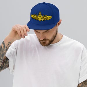 May include: A royal blue snapback cap featuring a gold embroidered United States Navy Surface Warfare insignia on the front panel. The hat has a structured crown, a flat brim, and a solid color design with detailed stitching on the emblem.
