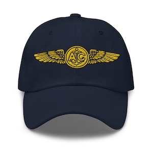 May include: A navy blue baseball cap with a gold embroidered emblem. The emblem features wings, an anchor, and the letters "AC". The cap has a curved peak and a structured crown. A classic, adjustable hat.