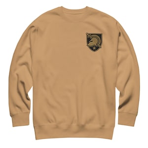 May include: A tan crewneck sweatshirt with a black and gold emblem on the upper left chest. The emblem features a helmet, sword, and star design. The sweatshirt is made of a soft material.