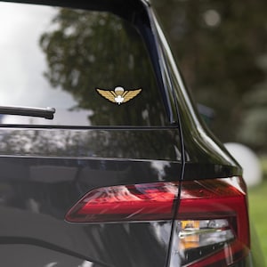 May include: A gold and black emblem featuring a parachute, wings, and a maple leaf is affixed to the rear window of a black vehicle. The emblem is a sticker, likely for a car or truck, and is visible against the backdrop of a blurred outdoor scene.