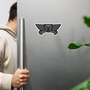 May include: A black and gray sticker featuring a stylized winged cougar design. The sticker has a white border and is affixed to a silver refrigerator door. The cougar has a fierce expression.