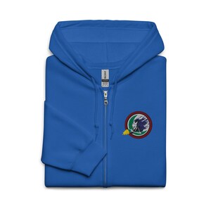 Puede incluir: Sudadera con capucha azul real con cremallera y un parche bordado circular. El parche presenta una ilustración en blanco y gris de una cabeza de dragón con un borde rojo, verde y amarillo. La sudadera tiene una cremallera completa.