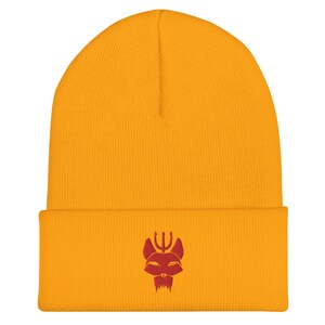 Puede incluir: Un gorro de punto amarillo con un logotipo rojo bordado de una cabeza de animal estilizada con un tridente en la parte superior.