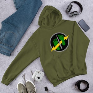 Pode incluir: Uma camisola de capuz verde com um gráfico circular preto e cinza. O gráfico apresenta um raio amarelo, duas pegadas verdes e uma faca cinzenta.
