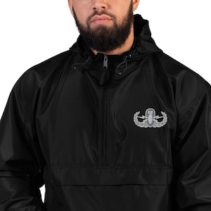 May include: A black windbreaker jacket featuring a quarter-zip front closure, a drawstring hood, and an embroidered silver-toned military-style EOD badge on the left chest. The jacket is constructed from a smooth, water-resistant material.