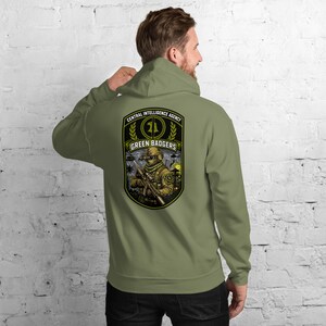 May include: Olive green hoodie with a graphic on the back featuring a soldier holding a rifle, the text "CENTRAL INTELLIGENCE AGENCY", and "GREEN BADGERS". The hoodie has a drawstring hood and a kangaroo pocket.