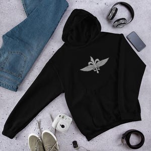 May include: Black hooded sweatshirt with a silver emblem on the chest. The emblem features a fleur-de-lis design with wings. The sweatshirt has a front pocket and a drawstring hood. The image also shows blue jeans, headphones, a phone, and a camera.