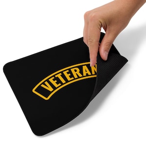May include: Black rectangular mousepad with the word "VETERAN" in yellow, arched across the top. A hand lifts the edge, showing a textured, black underside. The mousepad is approximately 25 cm long.
