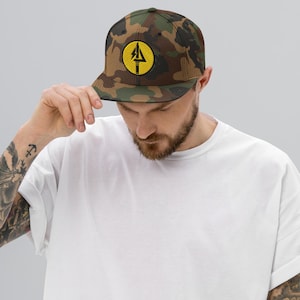 Könnte beinhalten: Eine Baseballkappe mit Camouflage-Muster und flachem Schirm. Die Kappe hat einen gelben runden Aufnäher mit einem schwarzen Grafikdesign. Eine Person mit einem weißen T-Shirt hält die Kappe.