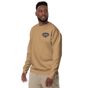 May include: A tan sweatshirt with a small embroidered emblem on the upper left chest. The emblem features a design with wings and the text "LAND-SOAR". The sweatshirt has long sleeves and a crew neck.