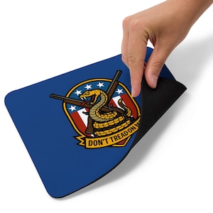 May include: A blue mousepad featuring a coiled yellow snake with a red tongue, crossed rifles, and the text "DON'T TREAD ON ME." The design is set against a shield with stars and stripes.