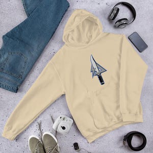 May include: A cream-colored hoodie with a front pocket and a hood. The hoodie features a graphic of a stylized arrow with a knife blade. The image also shows blue jeans, sneakers, headphones, a phone, a camera, and a belt.