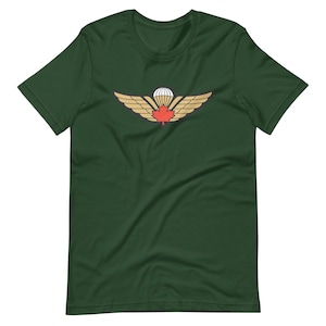 Könnte beinhalten: Dunkelgrünes T-Shirt mit einem goldenen und roten Emblem. Das Emblem zeigt ein Ahornblatt, Flügel und einen Fallschirm. Das T-Shirt besteht aus einem weichen Material.