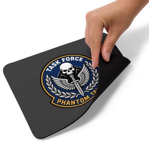 Può includere: Tappetino per mouse rettangolare nero con un emblema circolare. L'emblema presenta le parole "TASK FORCE" e "PHANTOM TA", un teschio, ali, una spada e foglie di alloro, in blu, bianco e oro.