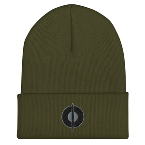 Pode incluir: Gorrinho verde-oliva com um emblema bordado em preto e cinza na frente. O emblema é um círculo com uma linha vertical que o atravessa. O gorro é feito de um material de malha macia e tem uma aba dobrada.
