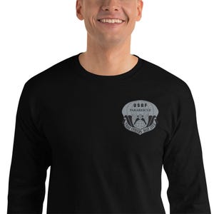 Puede incluir: Camiseta negra de manga larga con cuello redondo. La camiseta presenta un emblema gris en el pecho izquierdo con el texto "USAF Pararescue" sobre un diseño de paracaídas y las palabras "That Others May Live".