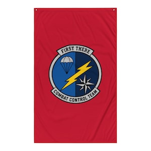 Op de afbeelding: Een rechthoekige rode vlag met een cirkelvormig embleem in het midden. Het embleem toont een blauwe achtergrond met een witte parachute, een gele bliksemschicht en een zwart-witte kompasster. De tekst FIRST THERE COMBAT CONTROL TEAM staat in het zwart op een witte rand.