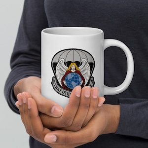 Può includere: Tazza in ceramica bianca con un emblema dettagliato raffigurante una figura alata che tiene in mano un globo, su uno sfondo a forma di paracadute. L'emblema include il testo "THAT OTHERS MAY LIVE". La tazza è tenuta con entrambe le mani.
