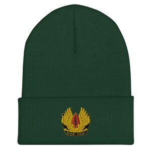 May include: A dark green knit beanie with a gold embroidered emblem featuring a red and yellow spear with wings. The emblem also includes the text "SINE PARI".