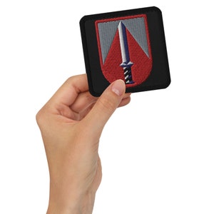 Op de afbeelding: Een zwarte geborduurde patch met een rood en grijs schild en een zilveren zwaard. Het zwaard heeft een blauw en wit handvat.