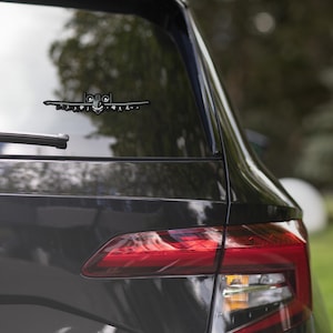 May include: A black car with a sticker of a military aircraft on the rear window. The sticker is a silhouette of the aircraft with a cartoon face.